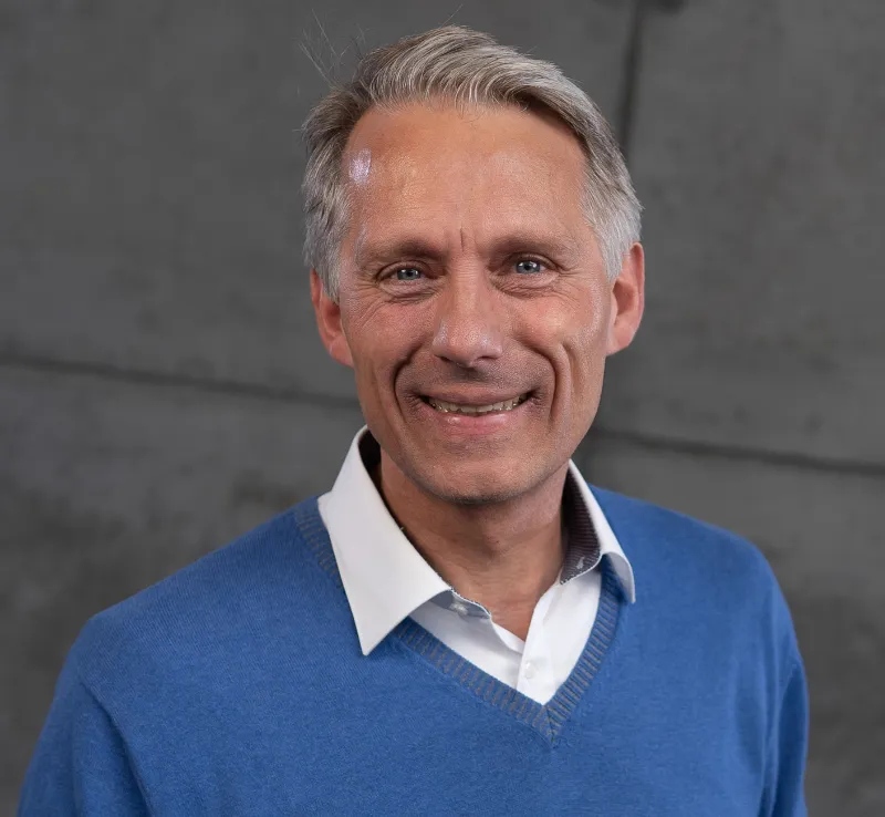 Sven Becker mit Hemd und blauem Pullover lächelt in die Kamera.